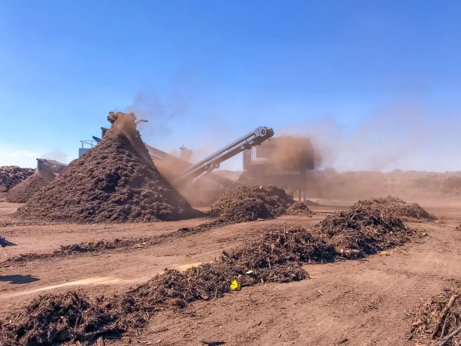 Compost windrows and a screening machine at a Rua-Con compost facility under a blue sky
